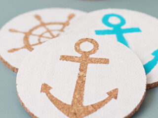 Three round coasters featuring nautical designs: a brown ships wheel, a brown anchor, and a blue anchor, all on a light background.