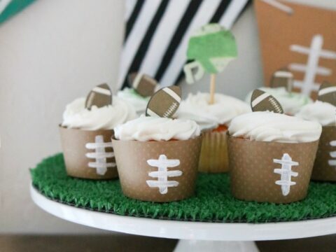 A cake stand holds football-themed cupcakes with brown wrappers designed like footballs. Above them are triangular banners in green, black-and-white stripes, and brown, including a 50 on the green one. The stand is covered with artificial grass.