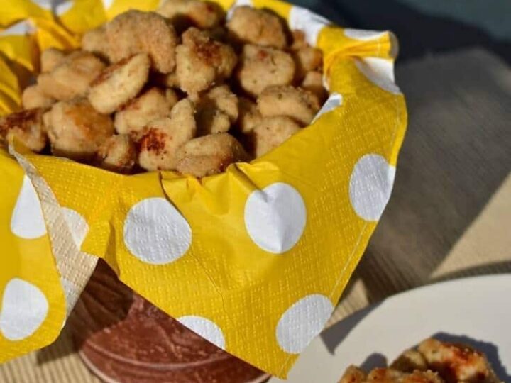 A basket lined with a yellow polka dot cloth is filled with crispy, golden-brown chicken nuggets. A white plate beside it holds a few more nuggets. Sunlight casts soft shadows on the arrangement.