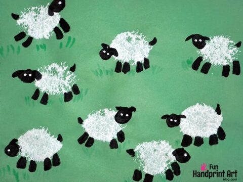 Childrens handprint art of fluffy white sheep with black faces and legs. The sheep are painted on a green background, scattered playfully across the image.