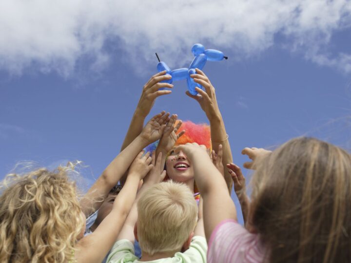 A group of children reaching up towards a person holding a blue balloon animal. The person is wearing a colorful clown wig, and the sky is bright and clear in the background. The scene exudes joy and excitement.