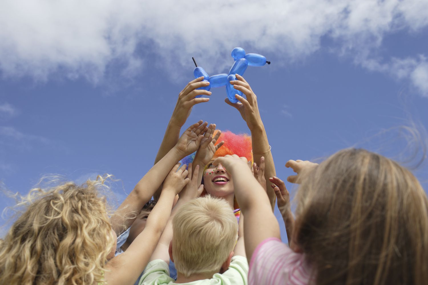 A group of children reaching up towards a person holding a blue balloon animal. The person is wearing a colorful clown wig, and the sky is bright and clear in the background. The scene exudes joy and excitement.