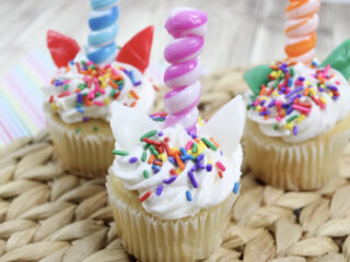 Three unicorn-themed cupcakes with swirled horn-shaped candles, colorful sprinkles, and white frosting are displayed on a woven mat. Each cupcake has candy ears and a striped candle, creating a whimsical look.