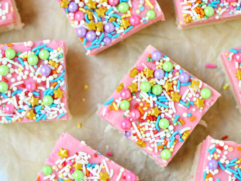 Colorful fudge squares topped with pink icing and decorated with colorful sprinkles and small sugar pearls on parchment paper.
