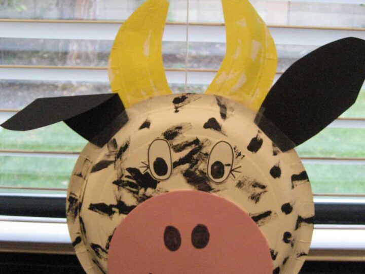 A craft cow face made from a paper plate, decorated with black spots, a pink snout, and googly eyes. Yellow construction paper horns and black paper ears are attached. The background shows partially open blinds.