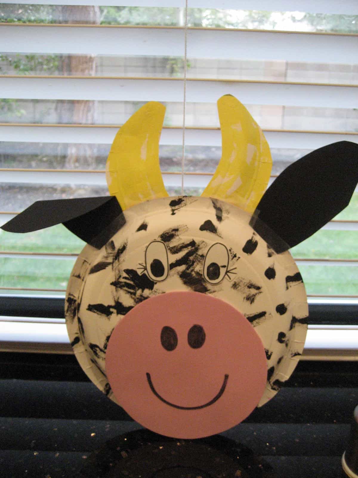 A craft cow face made from a paper plate, decorated with black spots, a pink snout, and googly eyes. Yellow construction paper horns and black paper ears are attached. The background shows partially open blinds.