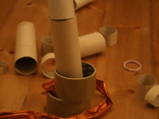 A wooden table with stacked and scattered cardboard tubes and rings. An orange ribbon is partially wrapped around one tube. A small hair tie lies next to them.