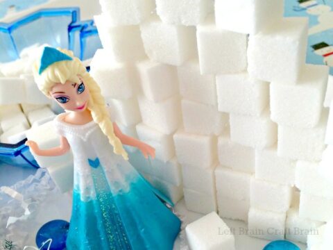 A small Elsa toy figure stands beside a stack of white sugar cubes arranged like a wall. Blue decorative stones are scattered around the base.