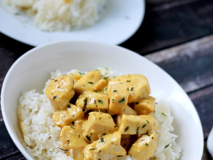 A bowl of white rice topped with creamy chicken pieces, garnished with herbs. Another plate with the same dish is set in the background on a dark wooden surface, alongside a fork and spoon.
