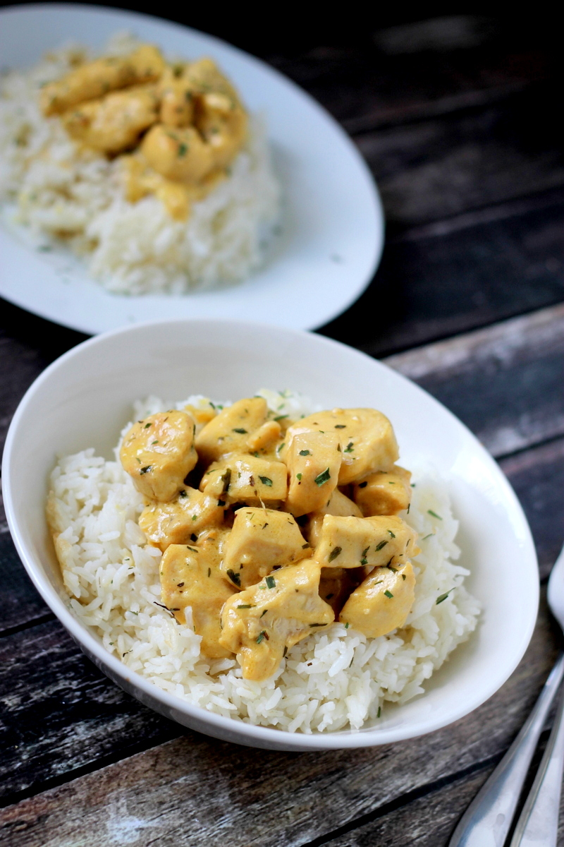 A bowl of white rice topped with creamy chicken pieces, garnished with herbs. Another plate with the same dish is set in the background on a dark wooden surface, alongside a fork and spoon.