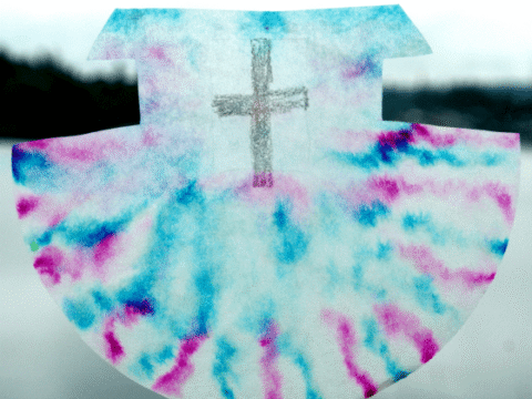 A colorful coffee filter craft with a tie-dye design in blue and pink features a cross drawn at the center. The background is blurry, and a logo reading meaningful mama is in the bottom right corner.