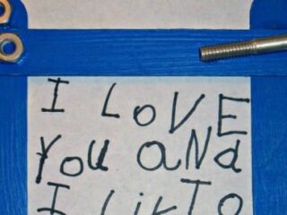 A blue popsicle stick picture frame decorated with two bolts and three nuts in the top left corner. Inside the frame, a handwritten note in black ink reads, I love you and I like you.