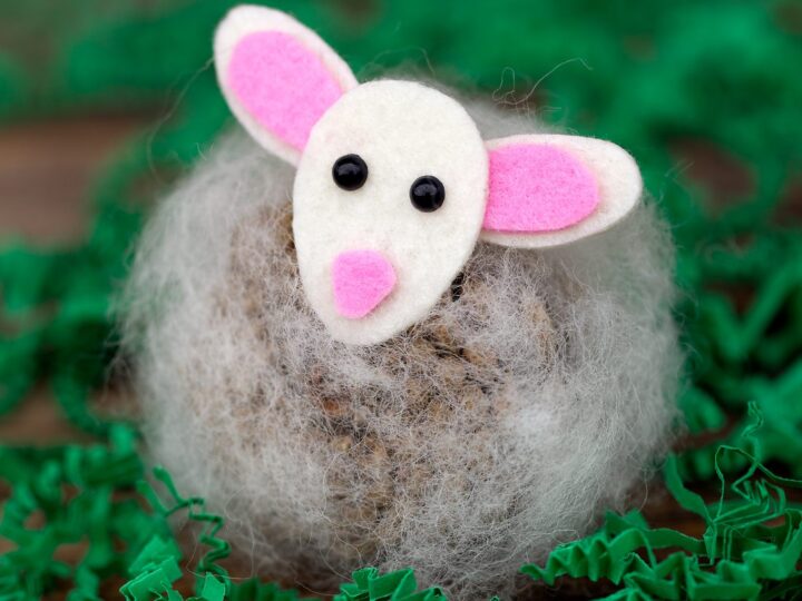 A cute mouse craft made from a stone, wrapped in fluffy material with a felt face and ears, lies on a wooden surface surrounded by green paper shreds.