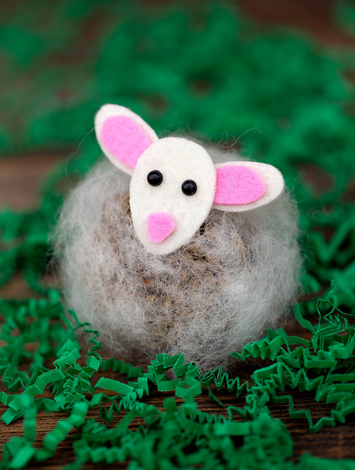A cute mouse craft made from a stone, wrapped in fluffy material with a felt face and ears, lies on a wooden surface surrounded by green paper shreds.