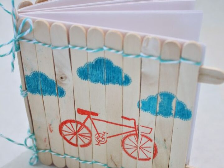 A handmade notebook with a cover made from wooden popsicle sticks. The front is decorated with blue cloud patterns and a red bicycle design. Bound with blue twine, the notebook has white pages inside.
