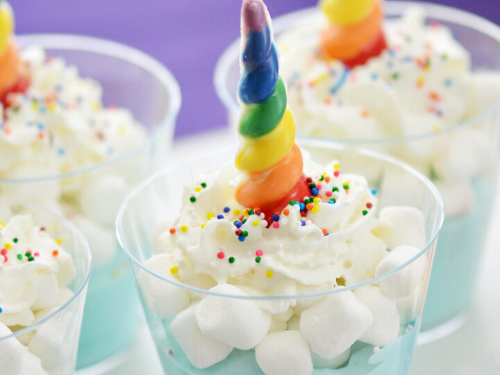 Three cups of blue pudding topped with whipped cream, colorful sprinkles, marshmallows, and a rainbow-colored candy shaped like a unicorn horn. The background shows a purple cloth and blurred colorful decoration.
