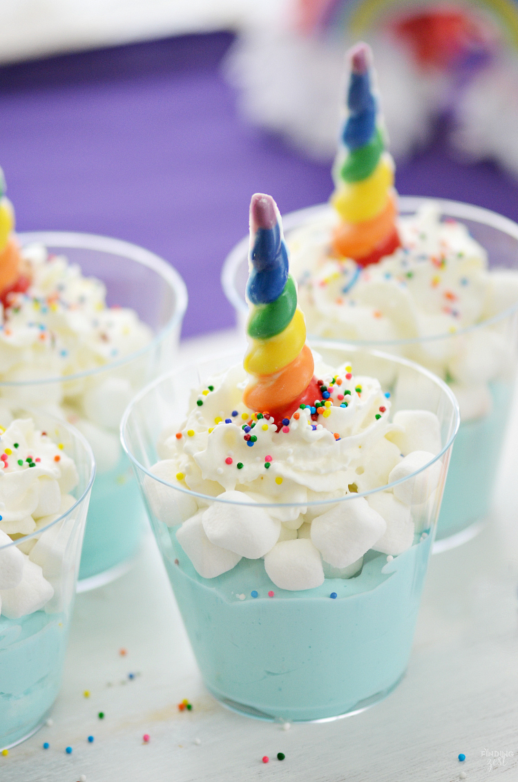 Three cups of blue pudding topped with whipped cream, colorful sprinkles, marshmallows, and a rainbow-colored candy shaped like a unicorn horn. The background shows a purple cloth and blurred colorful decoration.