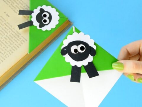 A green and white corner bookmark with a cute black and white sheep design is being placed on the corner of an open book. Another identical sheep bookmark is clipped to another page. Blue background.