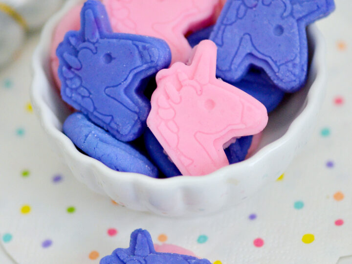 A white bowl filled with pink and purple unicorn-shaped candies sits on a white surface. The bowl is on a white napkin decorated with colorful polka dots. A silver-striped unicorn horn is in the background.