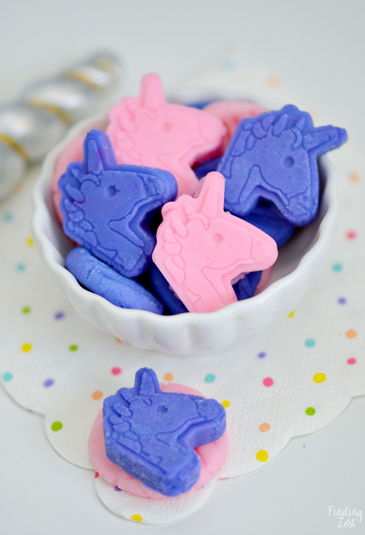 A white bowl filled with pink and purple unicorn-shaped candies sits on a white surface. The bowl is on a white napkin decorated with colorful polka dots. A silver-striped unicorn horn is in the background.