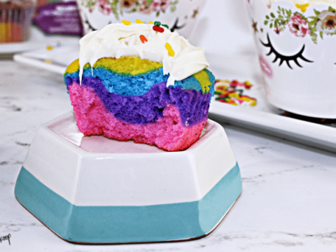 A colorful, half-eaten cupcake with layers of pink, purple, and blue, topped with white frosting and sprinkles. Its placed on a stacked hexagonal dish. In the background, theres a floral mug with eyelash designs and scattered sprinkles.