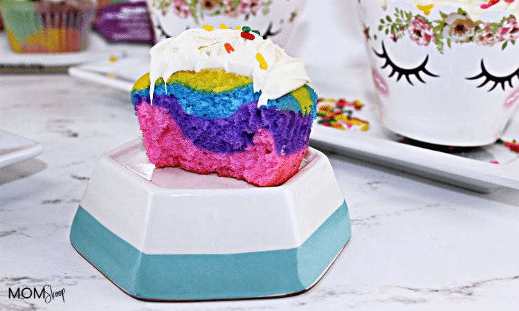 A colorful, half-eaten cupcake with layers of pink, purple, and blue, topped with white frosting and sprinkles. Its placed on a stacked hexagonal dish. In the background, theres a floral mug with eyelash designs and scattered sprinkles.
