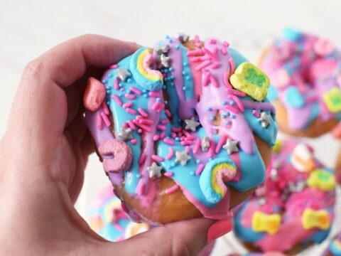 A hand holds a decorated donut with colorful pink and blue icing, topped with star-shaped sprinkles and various colorful cereal pieces shaped like stars, hearts, and rainbows.