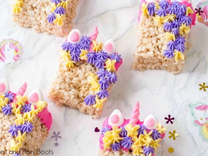 Colorful unicorn-themed Rice Krispie treats decorated with pastel purple, pink, and yellow frosting stars, and candy decorations resembling ears and a horn. They are placed on a decorative surface with unicorn-themed stickers.
