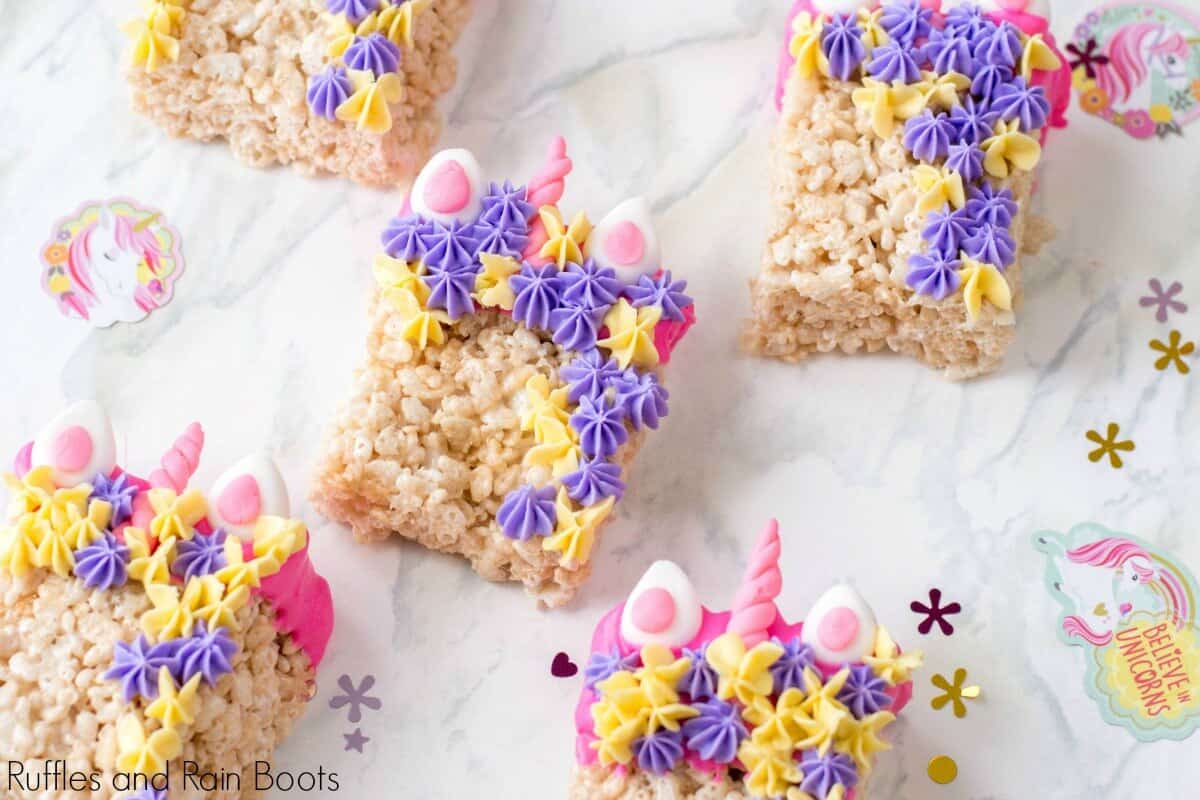 Colorful unicorn-themed Rice Krispie treats decorated with pastel purple, pink, and yellow frosting stars, and candy decorations resembling ears and a horn. They are placed on a decorative surface with unicorn-themed stickers.