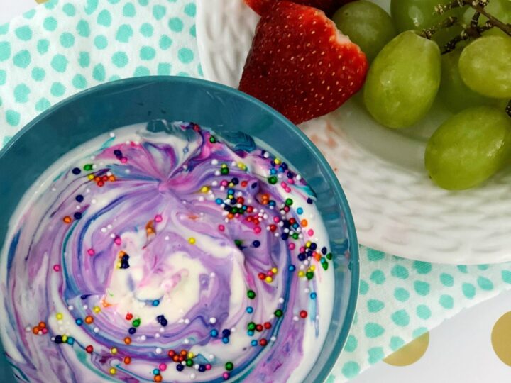 A blue bowl of swirled purple and white yogurt topped with colorful sprinkles is on a polka-dotted napkin. Beside it, a white plate contains green grapes and halved strawberries. The background is white with gold dots.
