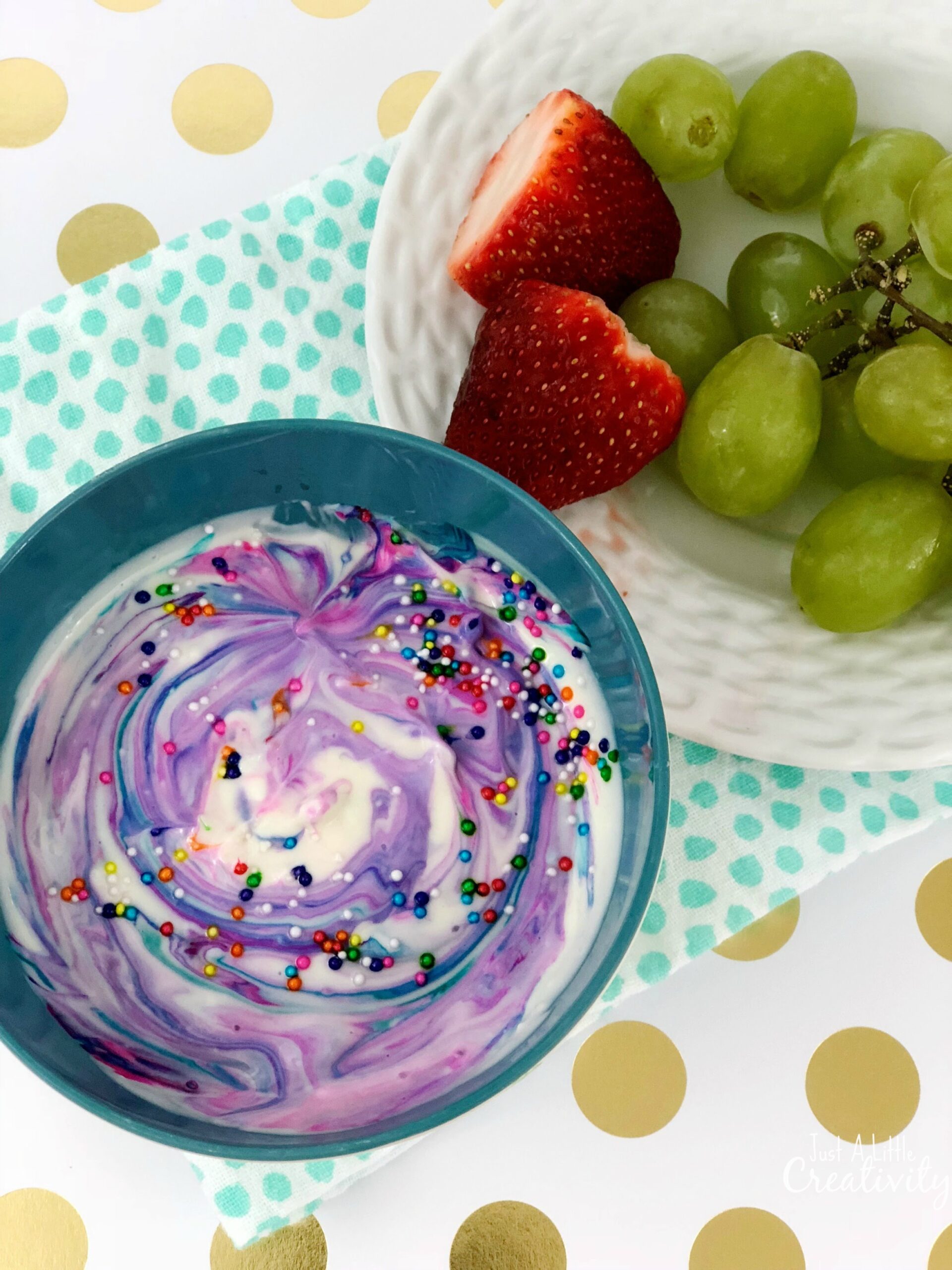 A blue bowl of swirled purple and white yogurt topped with colorful sprinkles is on a polka-dotted napkin. Beside it, a white plate contains green grapes and halved strawberries. The background is white with gold dots.