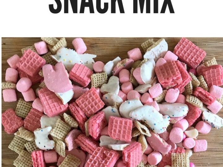 A heart-shaped pile of snack mix on a wooden surface. The mix includes pink wafer cookies, frosted animal crackers, cereal pieces, and pink mini marshmallows. Text above reads Unicorn Snack Mix.