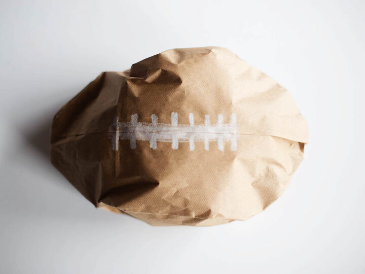 A crumpled brown paper bag shaped like a football with white stitches drawn on it, placed on a white surface.