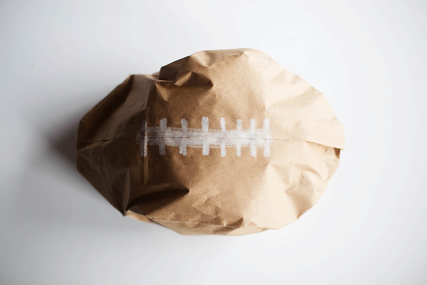 A crumpled brown paper bag shaped like a football with white stitches drawn on it, placed on a white surface.