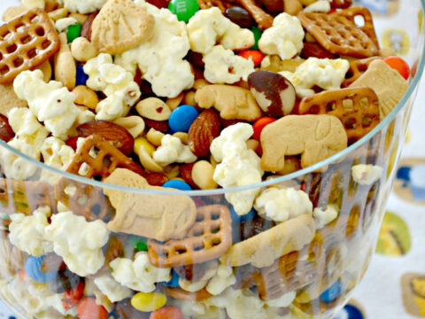 A glass bowl filled with a mix of popcorn, animal crackers, pretzels, chocolate candies, and nuts. The words Animal Cracker Popcorn Crunch are written above the bowl. The tablecloth features an animal pattern.