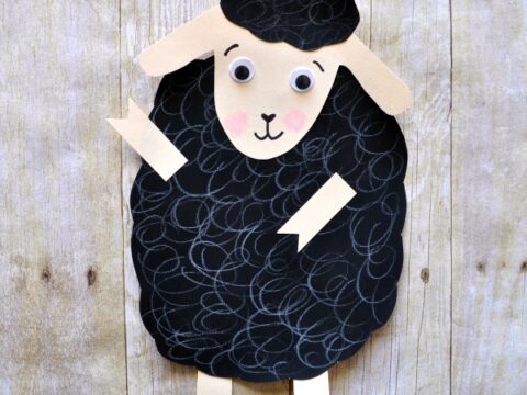 A craft of a black sheep made from paper against a wooden background. The sheep has googly eyes, rosy cheeks, and a smiling face, with a textured black body and beige paper limbs and ears.