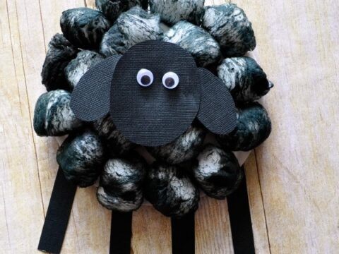 Craft project of a sheep made with black painted cotton balls and a paper head with googly eyes and ears. The sheeps legs are black paper strips. The background is a light wooden surface.