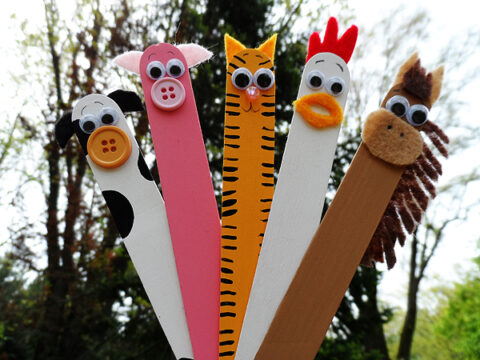 Crafted popsicle sticks decorated as animals stand upright: a cow with a button nose, a pink pig, a tiger with stripes, a chicken with a red comb, and a horse with a fuzzy mane. They are set against a blurred outdoor background.