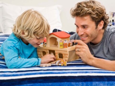 A smiling adult and child are lying on a bed playing with a wooden toy. The toy resembles a house or ark with small figures and animals. The adult has curly hair and is wearing a striped shirt, while the child is wearing a blue long-sleeve shirt.