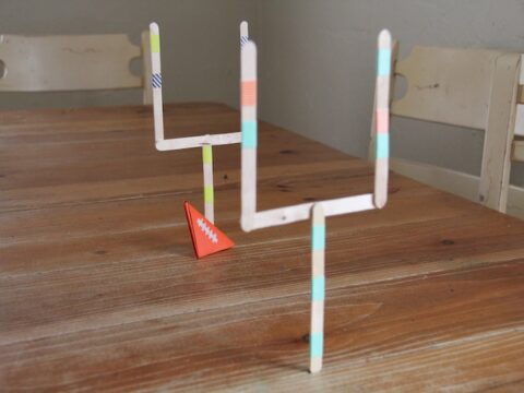 Two homemade goalposts made from popsicle sticks with colorful tape stand on a wooden table. A small, orange triangular football with a white lacing pattern is placed in front of one goalpost. Two white chairs are visible in the background.