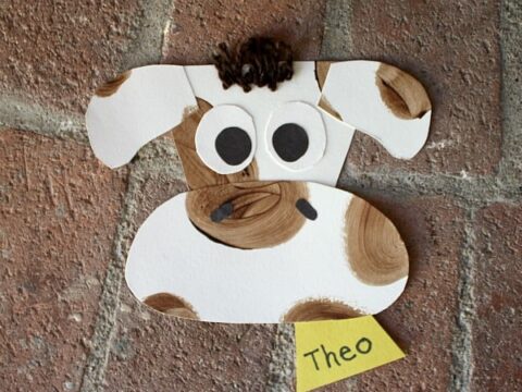 A paper craft of a cows face with brown spots on a brick background. The cow has large eyes and a small tuft of dark hair. A yellow tag with the name Theo is attached below.