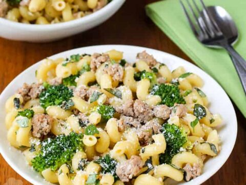A plate of creamy pasta with broccoli, crumbled sausage, and grated cheese. A green napkin with a fork and spoon is beside the plate. Another bowl of the same dish is partially visible in the background on a wooden table.