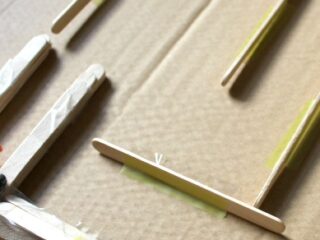 Close-up of a cardboard surface with wooden craft sticks arranged in parallel lines. A small toy car is attached to one of the sticks using tape. The arrangement suggests a DIY project or homemade track design.