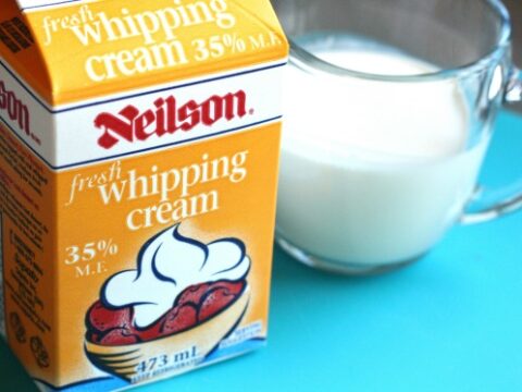 Carton of Neilson fresh whipping cream with 35% milk fat shown next to a glass mug filled with cream. The carton displays an image of cream topping a dessert. Both items are placed on a blue surface.