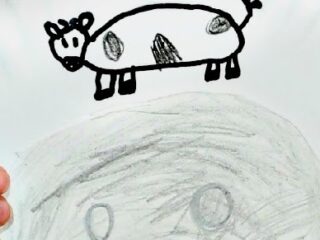 A child holds a paper plate with a drawing of a cartoon cow and simplistic facial features. The cow is sketched with black ink, standing atop a large grey circle, and has a spotted body and a tail.