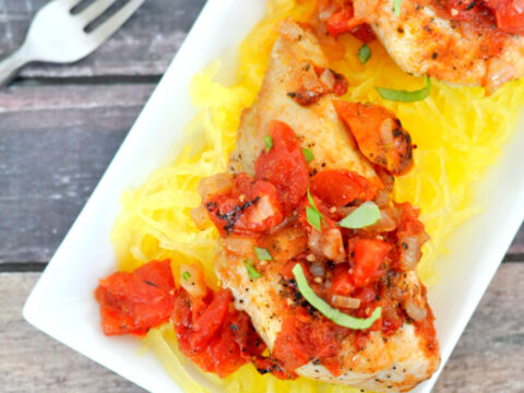 A plate of roasted chicken topped with diced tomatoes and herbs, served on a bed of spaghetti squash. A fork is placed beside the rectangular white plate on a wooden table surface.