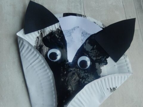 A paper plate craft shaped and painted to look like a badgers face. It features black and white paint, paper ears, and googly eyes. The text reads, Paper Plate Badger.