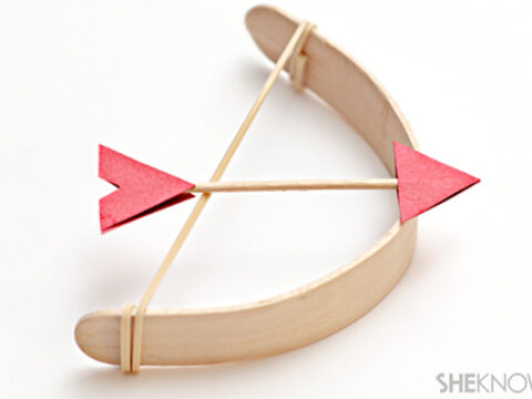 A small, handmade bow and arrow set crafted from popsicle sticks and rubber bands. The arrow features red paper fletching and the bow is curved with a rubber band string. The image has a white background and includes a watermark in the corner.
