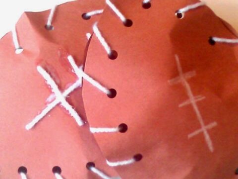 Two brown paper circles resemble footballs with white yarn laced through holes around the edges. White crayon markings mimic football stitching.