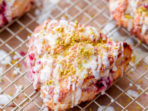 Close-up of freshly baked scones on a wire rack. The scones are drizzled with white icing and sprinkled with crushed pistachios, with visible berry pieces inside. The background shows more scones on a baking sheet.